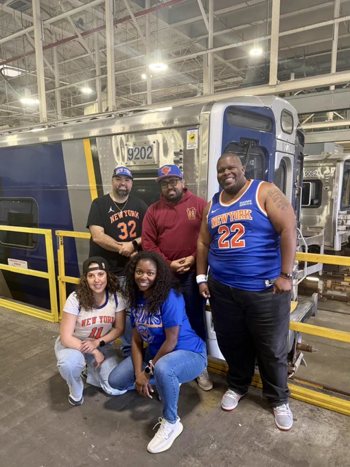 Five LIRR employees wearing Knicks merch, posed in front of an LIRR M9 train at the shop.