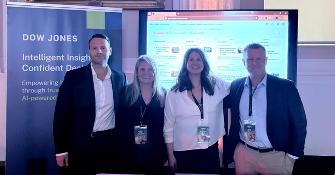 A group of four individuals stands in front of a Dow Jones display. They are inside a room with elegant decor, featuring a large screen showing a webpage. Each person is wearing a conference badge and smiling at the camera.