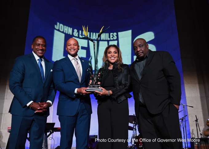 Four individuals standing together on a stage, smiling, and holding a large, decorative trophy. The backdrop features the name "John & Lillian Miles Lewis.