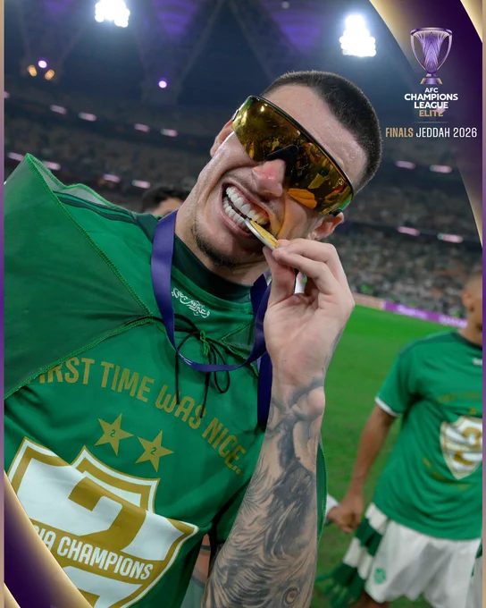 A joyful soccer player celebrates with a medal in his mouth after a victory at the AFC Champions League Elite Finals in Jeddah 2026. The scene is lively and filled with a sense of triumph.