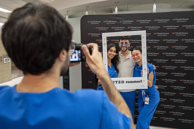 Three people pose together holding a large white rectangular frame decorated with small summer‑themed icons and the text “UTSD summer!” at the bottom. Two of them wear blue scrubs, and one is dressed in blue scrub pants and a shirt. A fourth person in the foreground takes their photo. Behind them is a backdrop repeating the UTHealth Houston School of Dentistry logo.