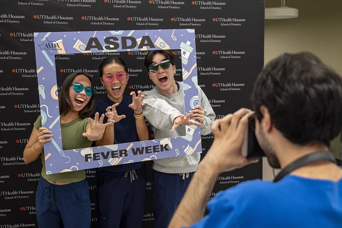 Three people pose enthusiastically while holding a large decorative frame labeled “ASDA FEVER WEEK,” illustrated with dental‑themed icons. They stand in front of a backdrop repeating the UTHealth Houston School of Dentistry logo. In the foreground, another person takes their photo with a smartphone.