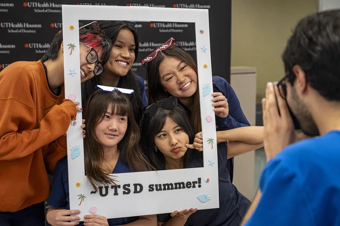 Five people pose together holding a large white rectangular frame decorated with small summer‑themed stickers like palm trees, seashells, and beach balls. The frame reads “UTSD summer!” A sixth person stands in front of them taking the photo. Behind the group is a black backdrop with the UTHealth Houston School of Dentistry logo.
