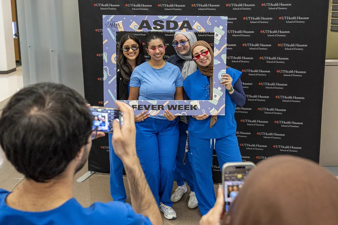 Four people wearing blue scrubs pose together holding a large decorative frame labeled “ASDA FEVER WEEK,” illustrated with dental‑themed icons. Behind them a black backdrop repeats the UTHealth Houston School of Dentistry logo. Two other people stand in the foreground taking photos with their phones.