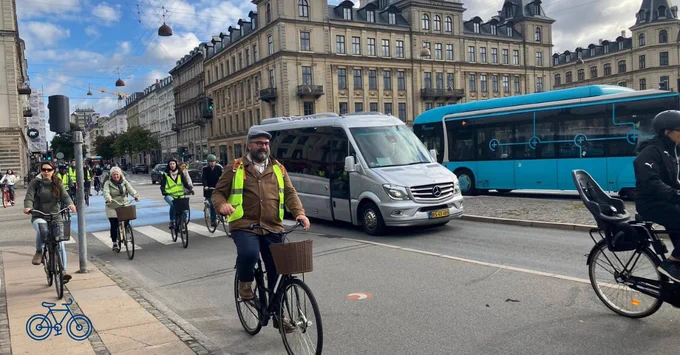 Group of cyclists riding on a city street with historic buildings and buses in the background under a partly cloudy sky.