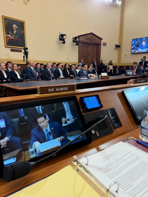 A photo from behind a committee dais desk that has a visual of Secretary Marco Rubio and a placard that reads "REP. McBRIDE".