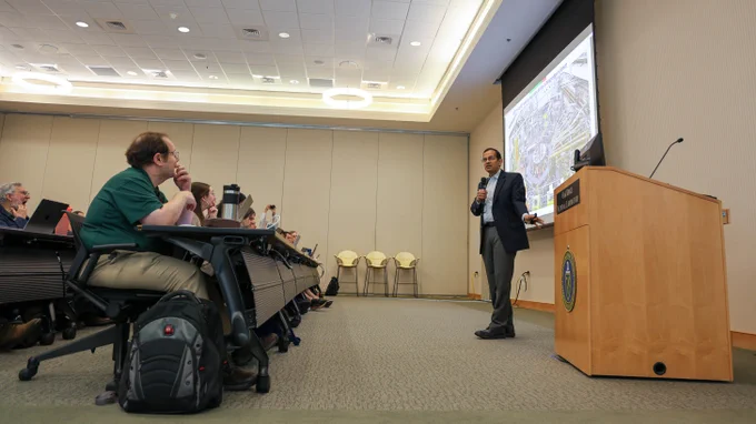 Arjun Shankar presents to attendees during the Oak Ridge Leadership Computing Facility User Summit, standing beside a large projection screen displaying information about advanced computing infrastructure and artificial intelligence-enabled research platforms.