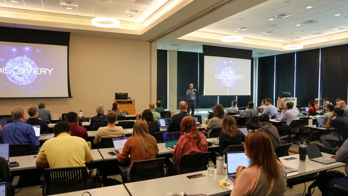 Matt Sieger addresses attendees during a session about the Discovery supercomputer at the Oak Ridge Leadership Computing Facility User Summit. Researchers seated throughout the room listen while viewing presentation slides displayed on large screens.