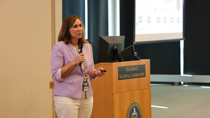 Ashley Barker speaks into a handheld microphone beside an Oak Ridge National Laboratory podium during the Oak Ridge Leadership Computing Facility User Summit.