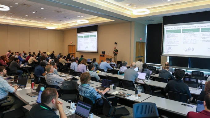 Josh Cunningham speaks into a handheld microphone while researchers and users follow along from conference tables equipped with laptops during the Oak Ridge Leadership Computing Facility User Summit.