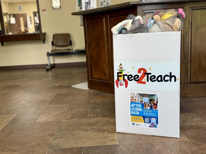 A large donation box labeled 'Free2Teach' is placed on the floor near a wooden counter in an indoor lobby area. The box is filled with various items in plastic bags, including stuffed animals. A flyer on the front of the box advertises an 'After School Bash' event with images of children and event details. In the background are chairs, a service window, and tan floor tiles.