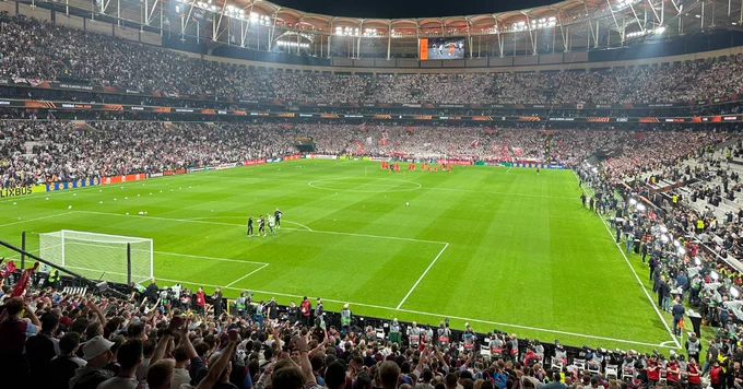 A high-angle night view of a massive football stadium filled to capacity under bright stadium lights. The green pitch is clearly visible from behind one of the goals, while the stands are densely packed with thousands of spectators wearing white.
