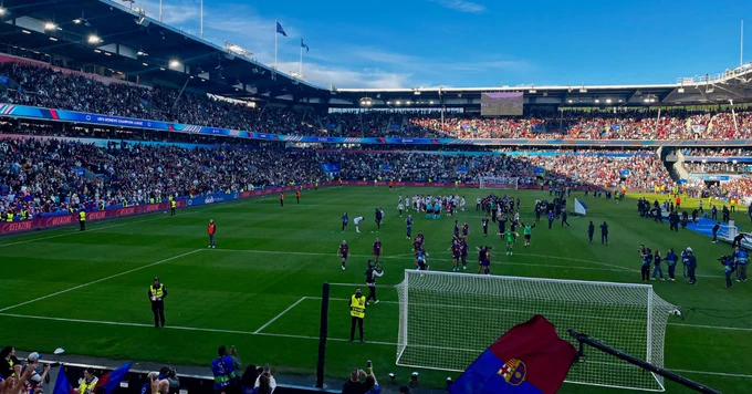 A high-angle view of a sunlit football stadium during a match event under a bright blue sky. Players and officials are scattered across the green pitch, with a white goal net visible in the foreground. In the bottom right corner, a fan waves a red and blue FC Barcelona flag.