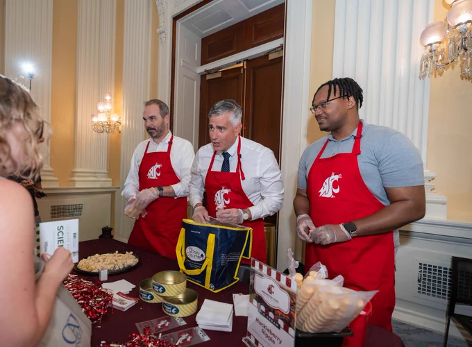 3 people stand behind a table wearing crimson aprons with a white WSU logo on the front. The person in the middle holds an ice cream cone and listens to a person on the other side of the table.