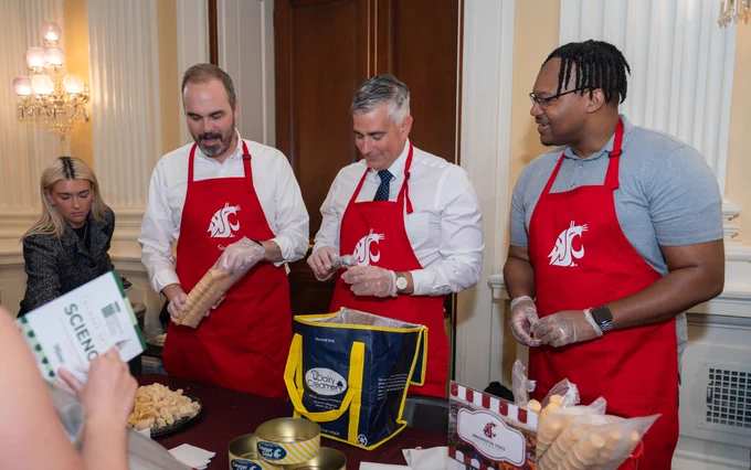 3 people stand behind a table wearing crimson aprons with a white WSU logo on the front. The person in the middle scoops ice cream into a cone, while the person on the left holds a bag of cones.