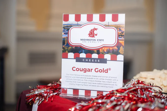 A sign with the WSU logo and an image of the WSU Pullman campus in the background. Words on sign: Washington State University cheese. Cougar Gold®. Our most famous and popular cheese and winner of several national and international awards! A rich, white cheddar with a smooth, firm texture. This unique cheddar has a depth and intensity that most people have never before experienced. Its creamy, lingering flavor will leave you wanting more!