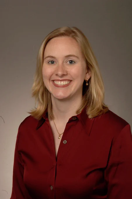 Person with shoulder-length blonde hair wearing a deep red button-up shirt and a delicate gold necklace against a plain background.