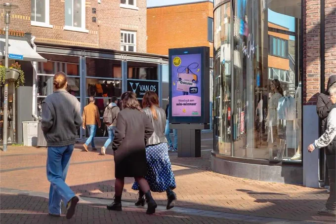 People walking on a sunny brick-paved street near a curved glass store window and a digital billboard with a lottery ad.