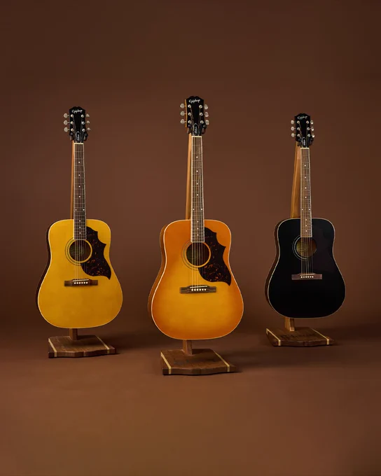 Three acoustic Epiphone Hummingbird Tribute  guitars are displayed on stands against a brown background. From left to right, the guitars are light wood, sunburst, and black in color.