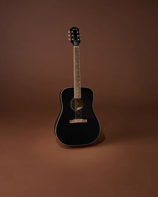 A black Epiphone Hummingbird Tribute acoustic guitar with six strings is standing upright. The background is a plain brown color.