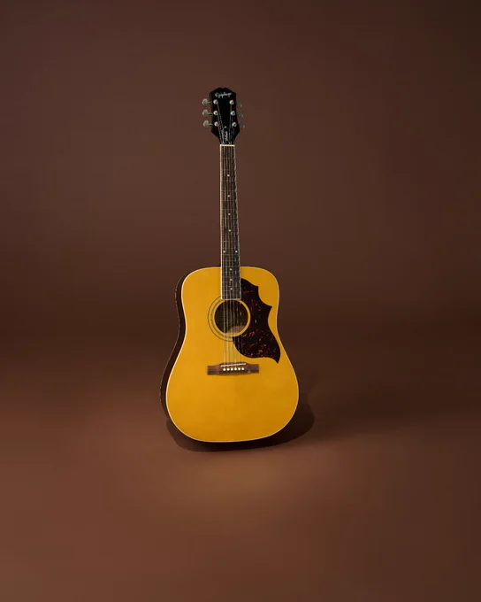 An Epiphone Hummingbird Tribute acoustic guitar with a natural wood finish is standing upright against a plain brown background. The headstock bears the Gibson logo.