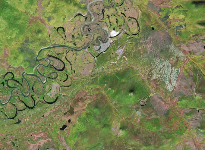 Satellite image of a region in Siberia, Russia, that includes the Batagaika Crater (lower right), which is manta-ray-shaped. To the northeast, a small hill overlooks the crater. The region is covered in greenery. The small village of Batagay is in the top middle, with the winding Yana River running from the lower left to above the town.