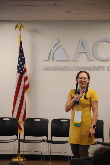 Dr, Chelsie Romulo smiles while holding a microphone and standing in front of the Arapahoe Community College logo.