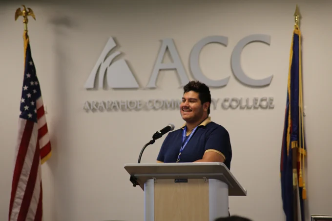 Tony Tasayco smiles behind a podium, speaking in front of an Arapahoe Community College logo.