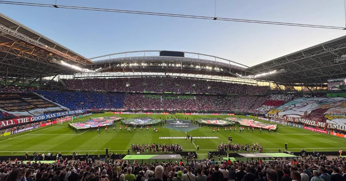 A wide, sweeping view of a stadium during a pre-match ceremony. Large banners are laid out on the center of the pitch. The grandstands are divided into massive fan displays, with a blue card stunt section on the left and a large illustrated tifo banner on the right.