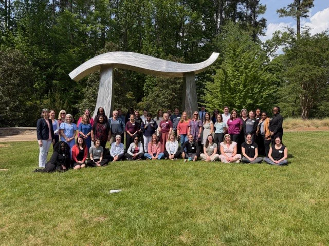 Large group of educators posing outdoors in front of a large pi sculpture on a sunny day.