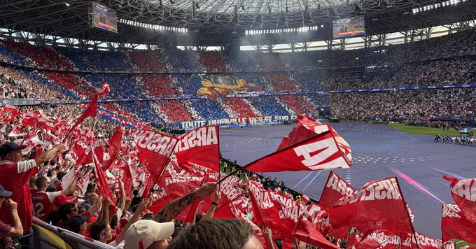 A view from the stands of a packed football stadium under a massive open-air roof structure. In the foreground, numerous fans wave large red and white flags bearing the word "Arsenal." In the distance, the opposing grandstand is covered in a massive red and blue mosaic display featuring a central illustration.