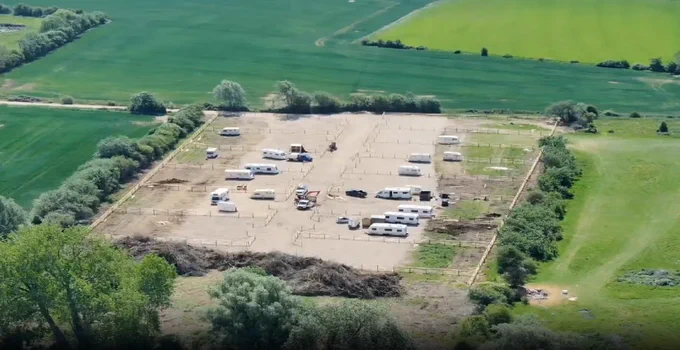Aerial view of a rural site with multiple static caravans and vehicles arranged within a large fenced area, surrounded by farmland and fields, with access tracks and hedgerows around the perimeter.