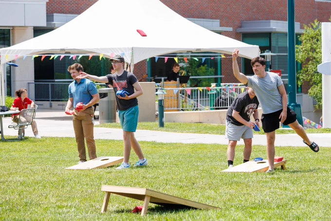 Employees play cornhole on a sunny campus lawn during an employee appreciation event, with tents and festive decorations in the background.