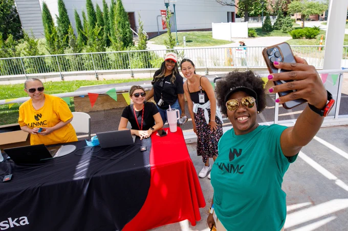 A group of university employees smiles for a selfie at a registration table during an outdoor employee appreciation event.