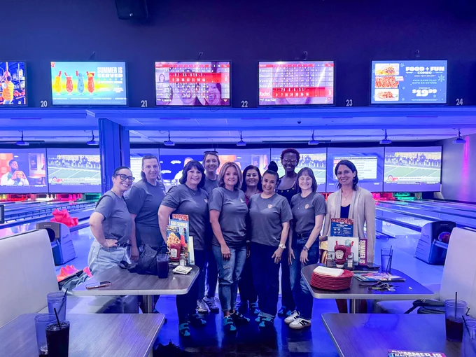 Ten SPPH administrative team members stand together at a bowling alley, with bowling lanes, score screens, tables, and blue and purple lane lighting in the background.