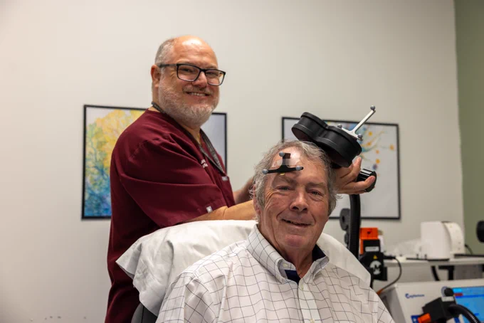 Joao L. de Quevedo, MD, PhD, prepares to administer Stanford Accelerated Intelligent Neuromodulation Therapy (SAINT) to Bill Henderson at UTHealth Houston. Henderson sits in a treatment chair wearing positioning markers while de Quevedo holds the transcranial magnetic stimulation device used to deliver targeted magnetic pulses.