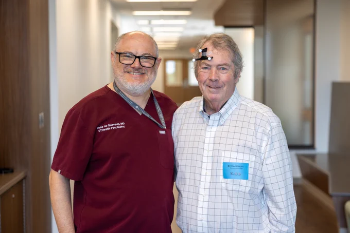 Joao L. de Quevedo, MD, PhD, poses with Bill Henderson at the Center for Interventional Psychiatry at UTHealth Houston. Henderson, the first patient in Texas to receive Stanford Accelerated Intelligent Neuromodulation Therapy (SAINT), smiles beside de Quevedo while wearing the positioning markers used during treatment.