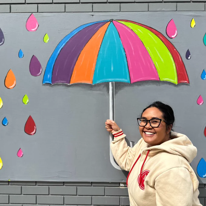 A person stands smiling next to a colorful mural of an umbrella and raindrops painted on a gray brick wall. The umbrella features vibrant sections in blue, orange, pink, green, and purple, with multicolored raindrops around it. The person wears glasses and a light-colored hoodie.