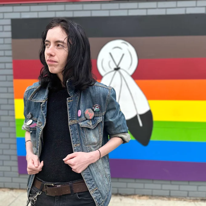 A person with long hair is standing in front of a mural featuring a stylized feather design against a backdrop of rainbow stripes. They are wearing a denim jacket with various pins. The setting appears to be outdoors with a paved surface.