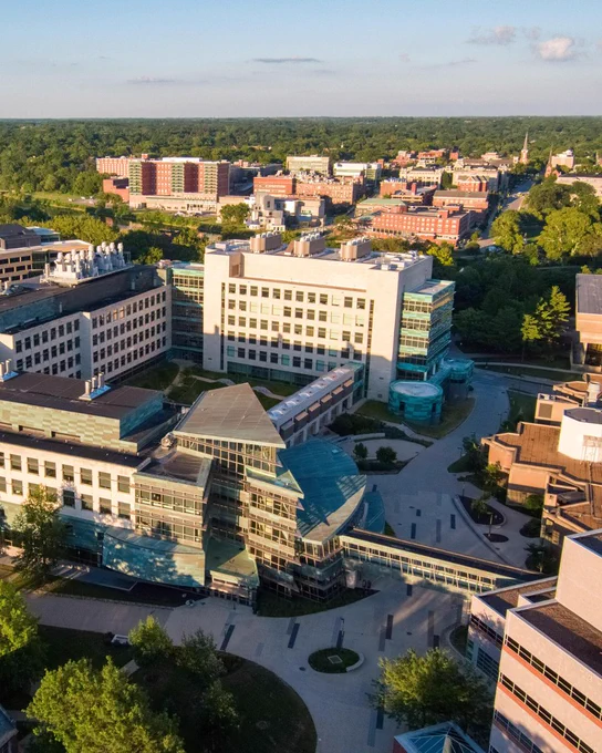 Aerial view of the University of Iowa Health Care campus in Iowa City during golden hour, with multiple hospital and medical research buildings connected by skywalks and courtyards. Trees and the surrounding city stretch into the distance under a clear evening sky.