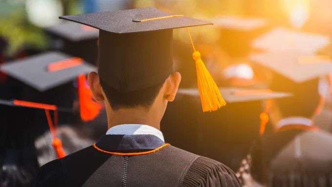 A person wearing a graduation cap and gown is seen from behind, standing among a crowd of graduates under bright sunlight.