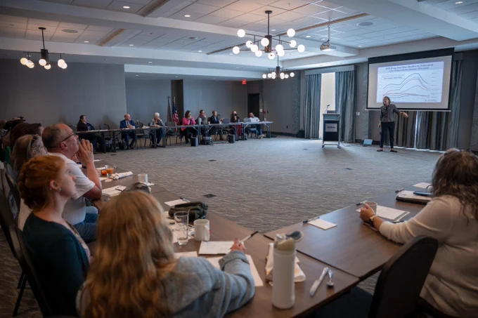 A presenter stands in front of a data slide on a large screen with a group seated around a U-shaped conference table. 