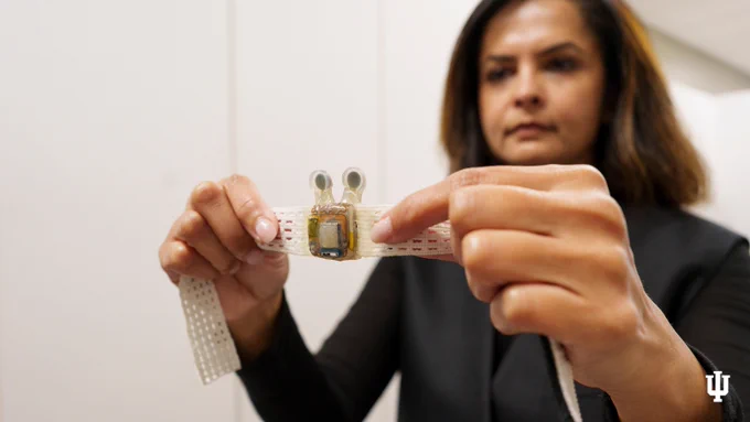 Rita Patel is holding up a piece of technology attached to a flexible band. The background is a plain indoor setting, and the Indiana University logo is in the lower right corner.