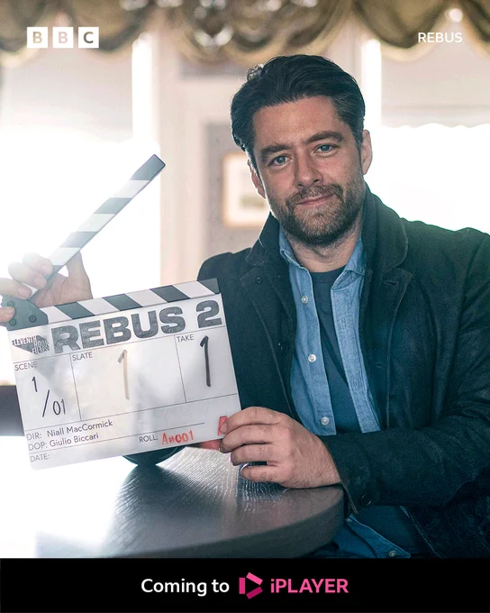 Richard Rankin as Rebus. He sits at a table on set of the drama and holds a clapperboard. Writing on the clapperboard reads: “Rebus 2, scene 1, slate 1, take 1.” 