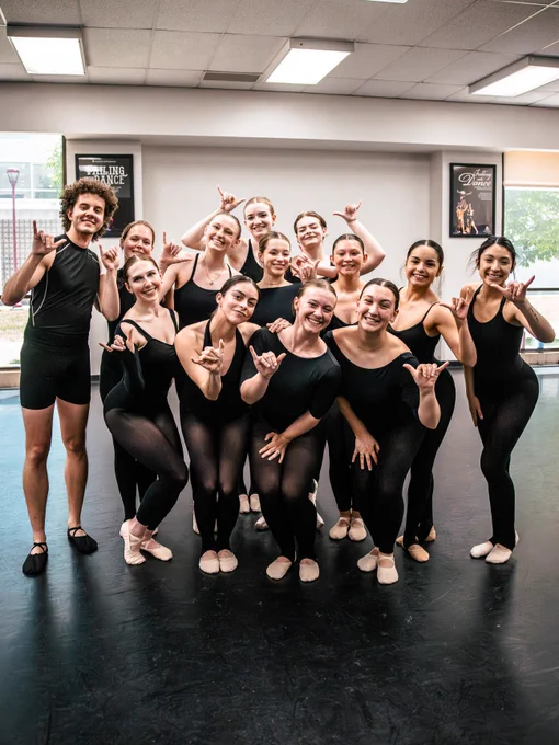 A group of ballet dancers pose together in a dance studio, each with one hand extended making a go buffs gesture. They are wearing black dance attire and ballet shoes. The studio has posters on the walls, one mentioning "The Studio" and another with a picture of a dancer.
