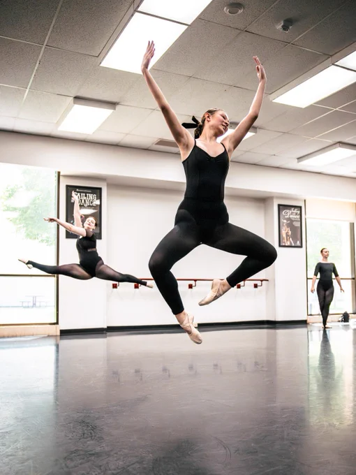 Ballet dancers practicing in a studio, one in mid-air with others engaged in various poses. Wall posters are on the wall in the background hanging above a bar mounted to a wall. 