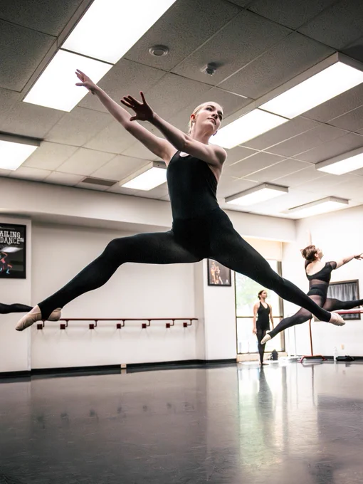 A ballet dancer in a black leotard and tights performs a grand jeté in a dance studio. Two others are practicing in the background, and the room is bright with natural light coming through the windows.