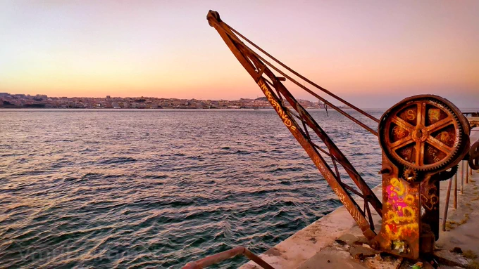 Grua metálica junto ao rio Tejo, em Cacilhas, ao pôr do sol, com reflexos na água e o horizonte iluminado.
