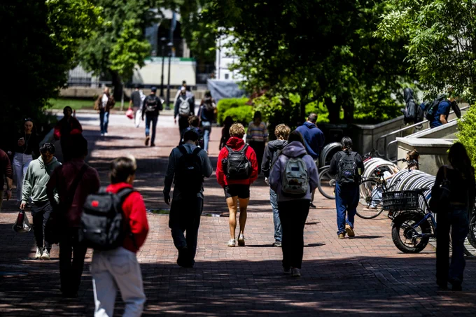 A busy outdoor scene with many people walking on a sunlit brick pathway. Most individuals wear casual clothes and carry backpacks. Lush green trees frame the path, with a building partially visible in the distance. Several bicycles are parked near a multi-tiered concrete structure on the right.