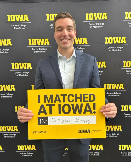 A person in a suit smiles while holding a yellow sign that reads "I MATCHED AT IOWA! IN: Orthopedic Surgery" in front of a backdrop with "IOWA Carver College of Medicine" repeated.